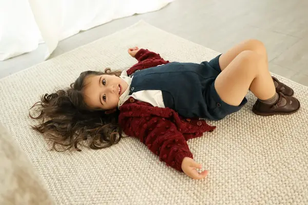 Toddler girl in a red cardigan lying on a cream rug, laughing, studio photography by Photoholic, Monsey NY